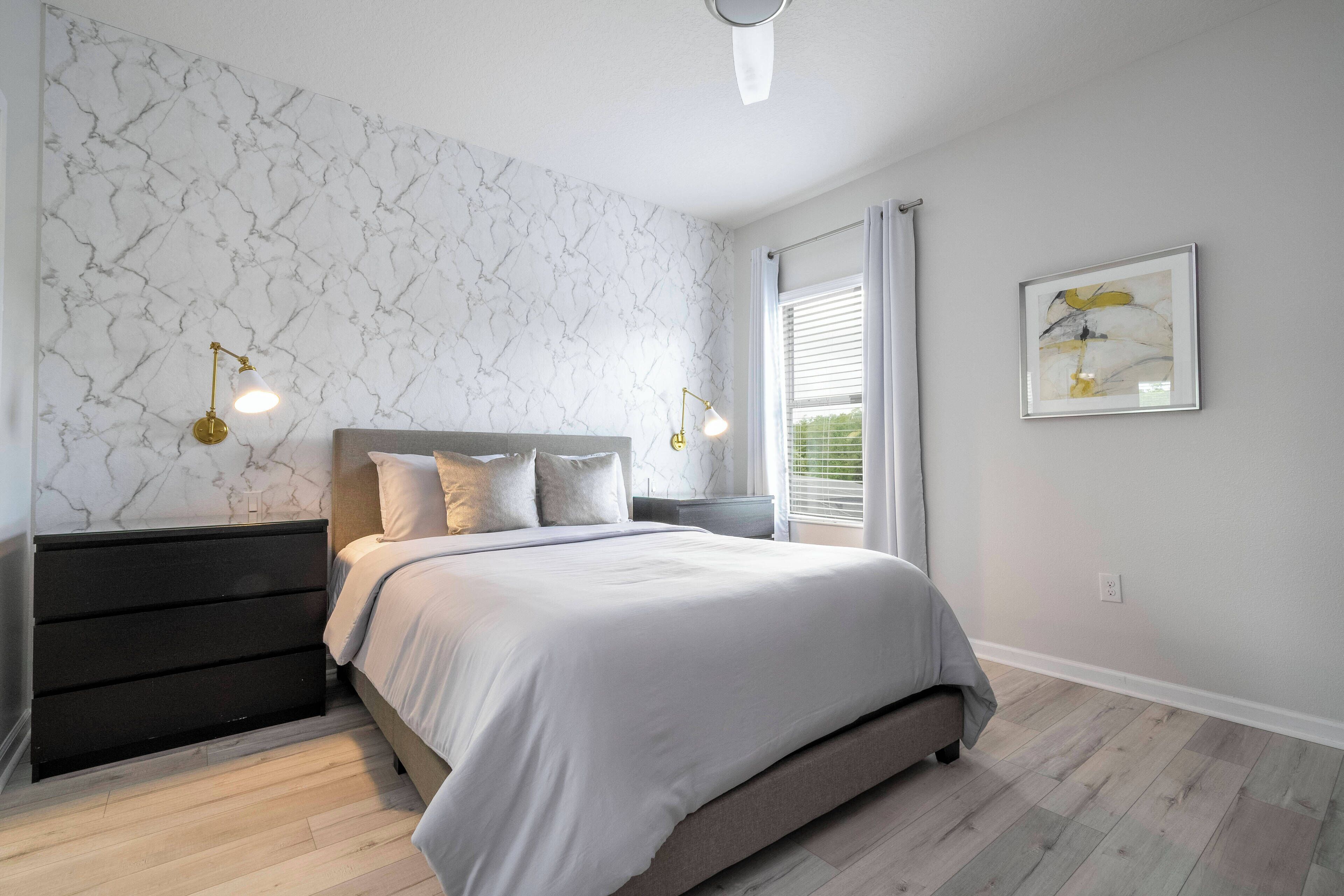 Een slaapkamer met een bed, nachtkastjes, een dressoir, behang met marmerprint, een plafondventilator en een raam met gordijnen.