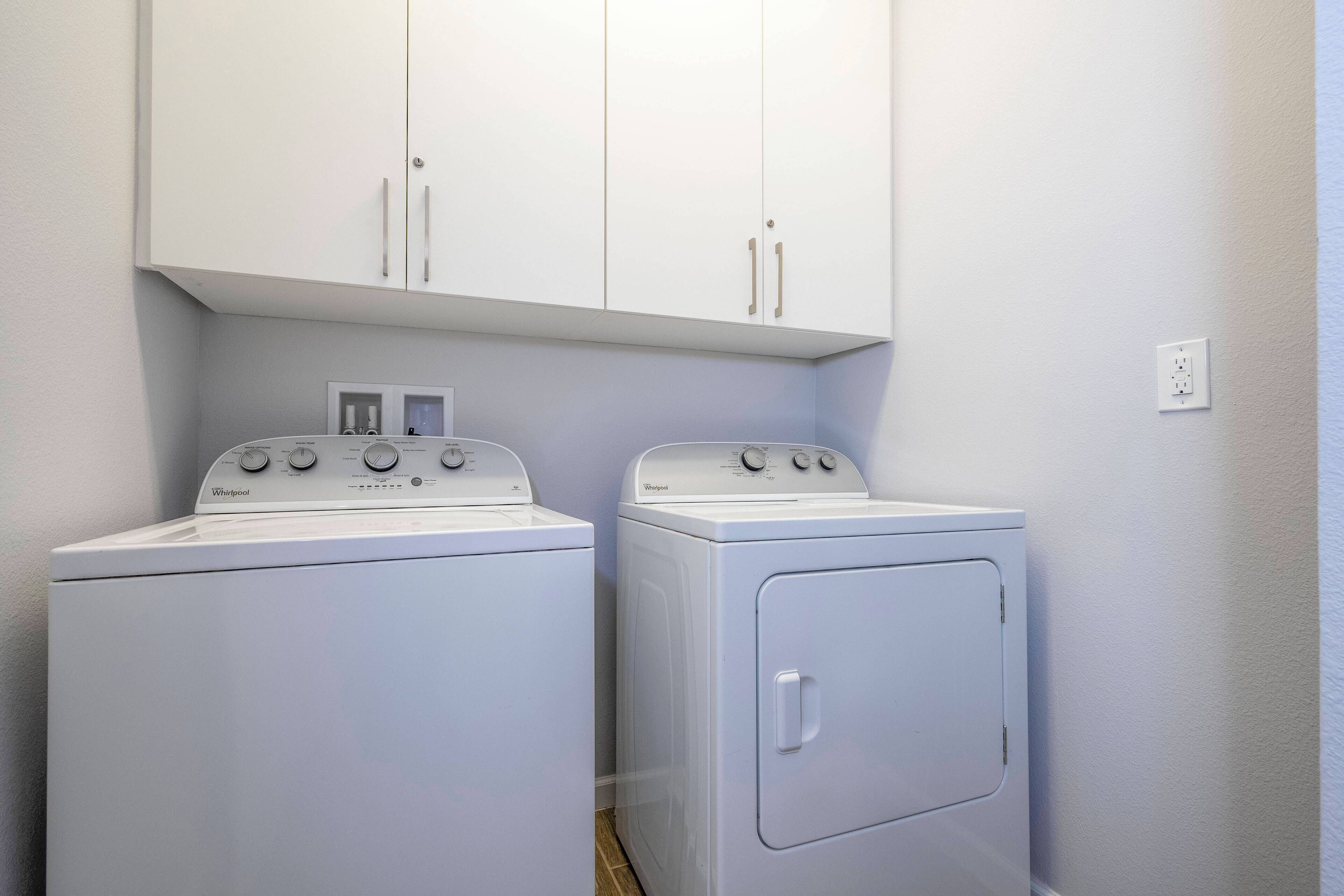 Pesutilassa on kaksi valkoista Whirlpool-merkkiä olevaa pesu- ja kuivauskonetta, niiden yläpuolella valkoinen kaappi ja seinässä valkoinen pistorasia.