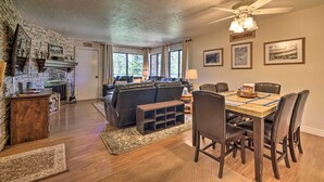 A spacious living room with a fireplace, a dining area with a wooden table and chairs, and a large window with curtains. - Slopeside Condo With Hot Tub + Game Room Access! (Brian Head)