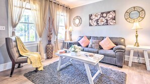 Un salon avec un canapé en cuir, une table basse et un fauteuil agrémenté d’un plaid jaune. On y trouve deux lampes sur des tables d’appoint, un miroir au mur et un tableau représentant des fleurs.