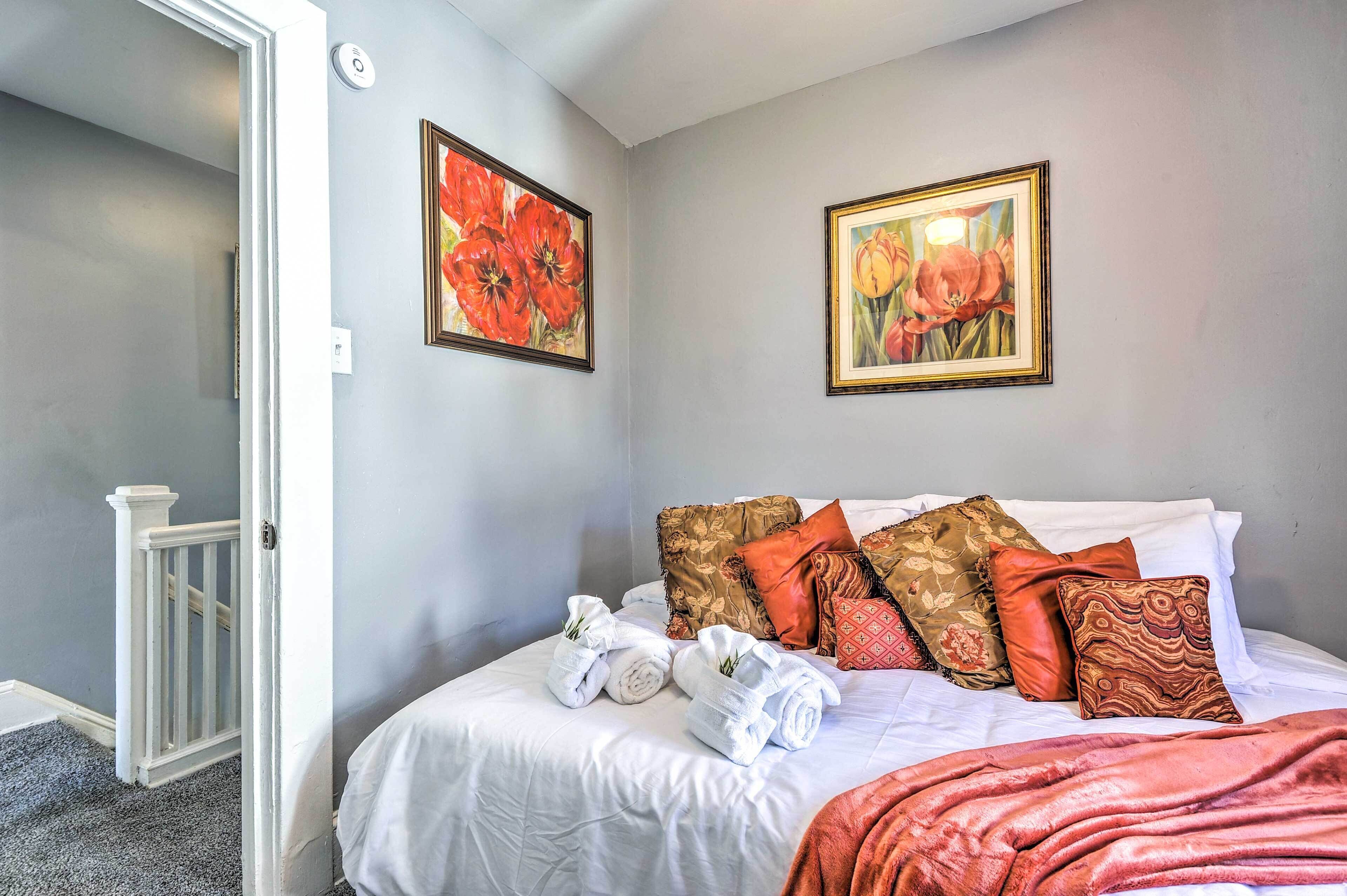 A neatly made bed with white linens, a red throw blanket, and several decorative pillows. There are two framed floral paintings on the wall above the bed.