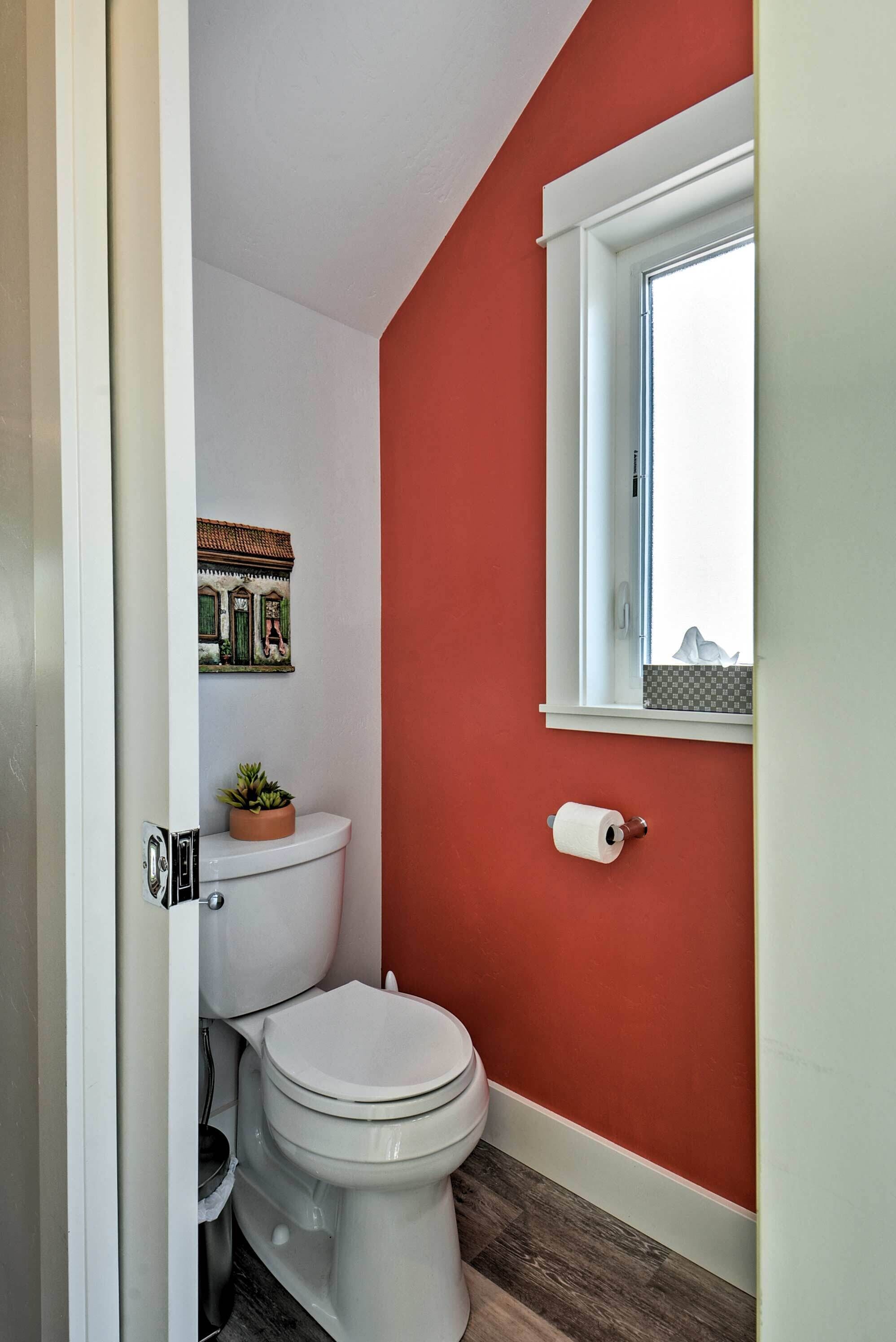 Un baño con una pared roja, un inodoro blanco, un ventanal pequeño y una planta sobre el tanque del inodoro.