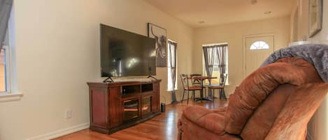 Una sala de estar con piso de madera, un sillón de cuero marrón, un televisor de pantalla plana sobre un soporte de madera, y una zona de comedor con una mesa y sillas.