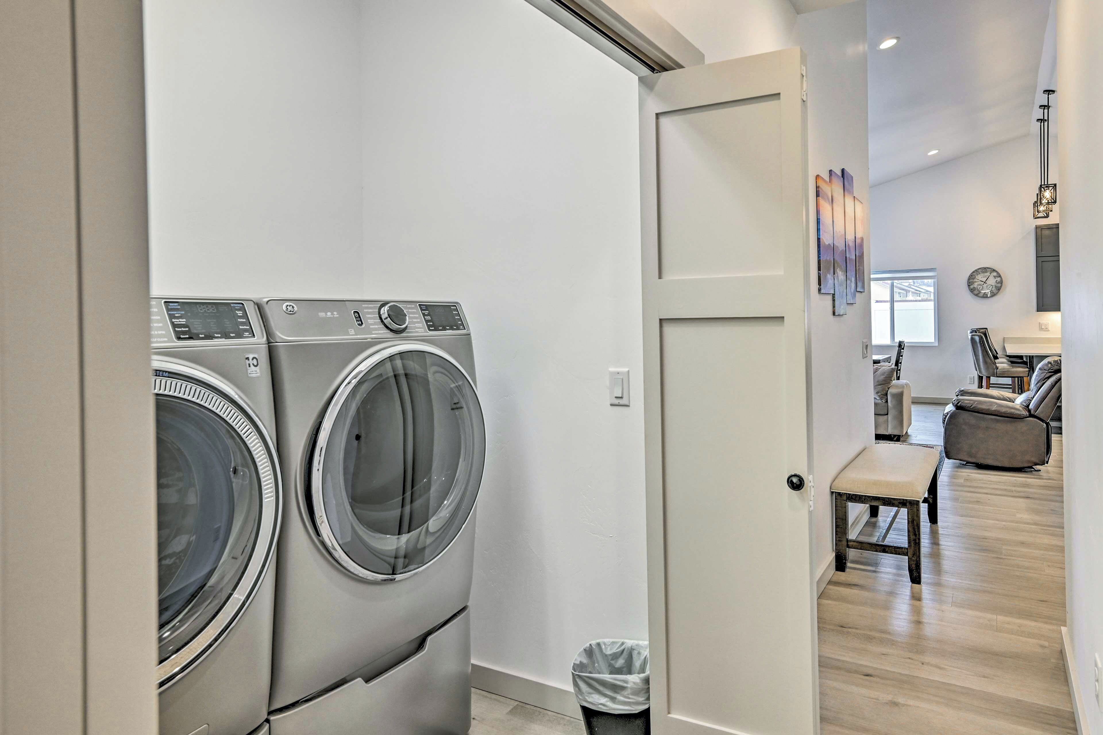 A laundry room with two front-loading washing machines, a trash can, and a door leading to a living area with a sofa and a coffee table.