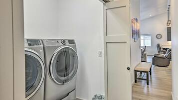 A laundry room with two front-loading washing machines, a trash can, and a door leading to a living area with a sofa and a coffee table.