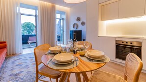 Un coin repas avec une table ronde en bois, dressée pour quatre personnes, entourée de chaises en osier. On aperçoit à l’arrière-plan une cuisine équipée d’un four moderne et d’un téléviseur.