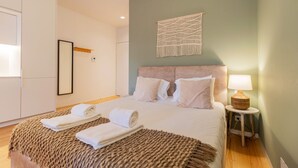 Un lit bien fait, avec deux serviettes enroulées, une tête de lit, une décoration murale en macramé, une lampe de chevet et une petite table.