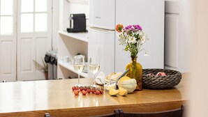 A wooden table with two glasses of white wine, a vase with flowers, a bowl of bread, and a plate with cheese and tomatoes.