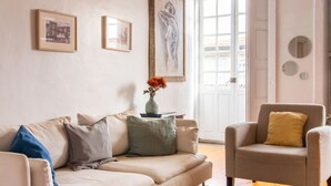 A living room with a beige sofa, a chair, and a vase with flowers. There are framed pictures on the wall and a door with a view of the outside.