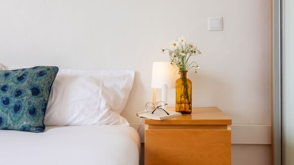 A neatly made bed with a white pillow and a blue patterned pillow, a bedside table with a lamp, glasses, and a vase with flowers.
