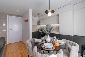 A modern dining area with a round table set for six, surrounded by grey upholstered chairs. A white vase with a feather arrangement and a glass pitcher of orange juice are on the table.