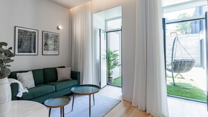 Un moderno salón con un sofá verde, dos mesas de centro de vidrio y un sillón colgante junto a un gran ventanal con cortinas blancas.