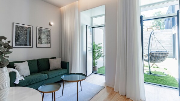 Ein modernes Wohnzimmer mit einem grünen Sofa, zwei Glastischen und einem Hängesessel neben einem großen Fenster mit weißen Vorhängen.