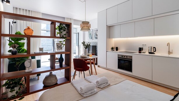 Una cocina moderna con armarios blancos, horno empotrado y fregadero. Hay una zona de comedor con una silla roja y una mesa. La habitación tiene un ventanal grande y un separador de madera con estantes que exhiben plantas y adornos.