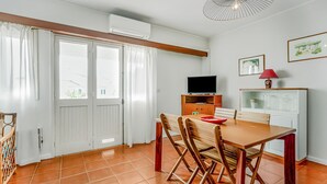 Un coin repas avec une table et des chaises en bois, un téléviseur posé sur un meuble et une fenêtre avec des rideaux.