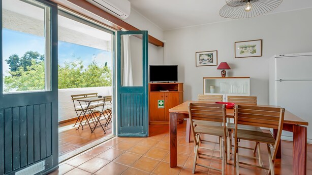 Un coin repas avec une table et des chaises, un téléviseur posé sur un meuble en bois, et une vue sur la verdure à l’extérieur.