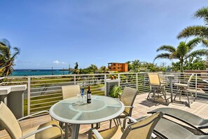 A terrace with a glass table, chairs, and a bottle of wine, overlooking a beach and ocean with palm trees and a building in the background. - Oceanside Vega Baja Beach House Paradise w/ Views! (Vega Baja)
