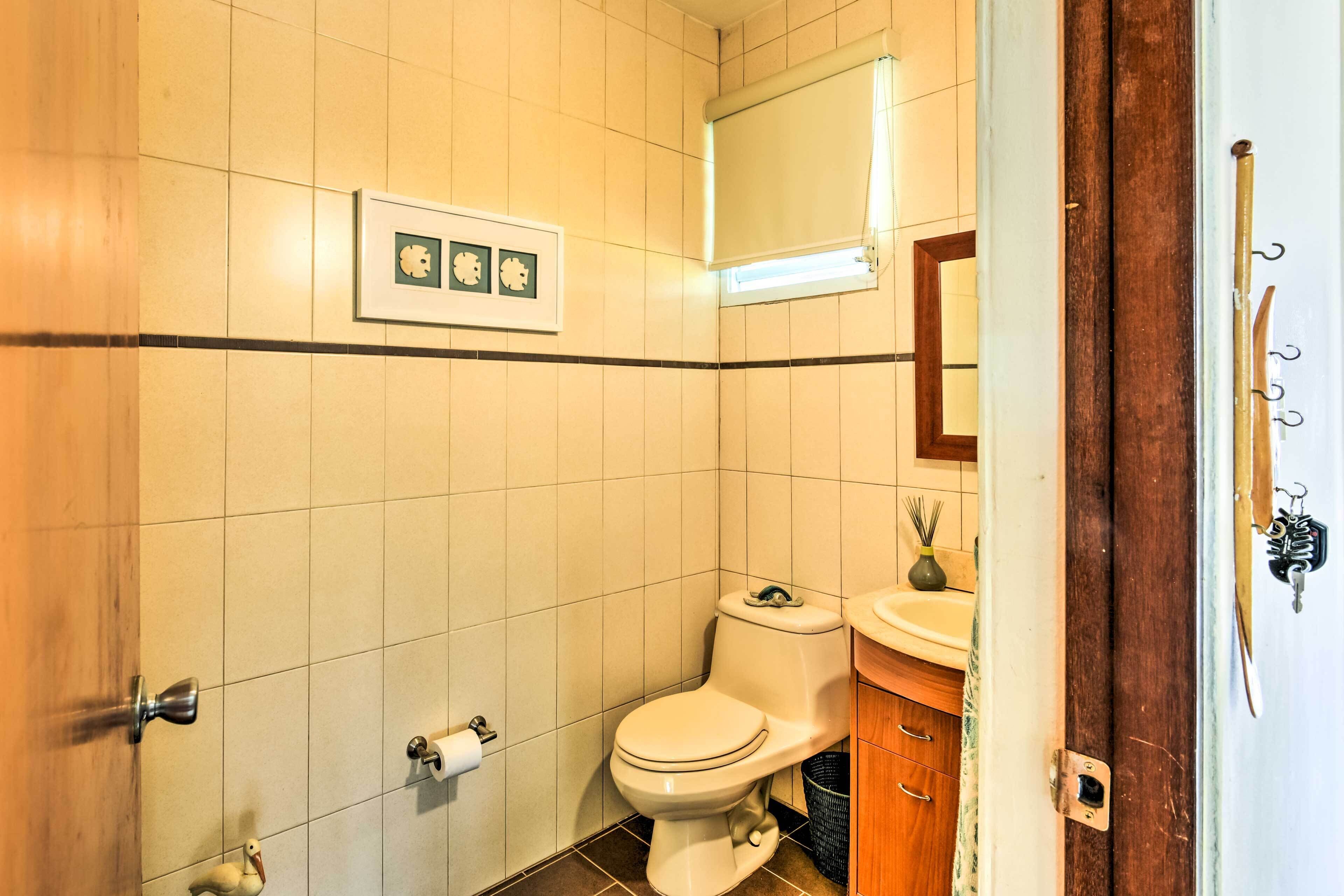A bathroom with a toilet, sink, and mirror. There is a framed picture on the wall and a window with blinds.