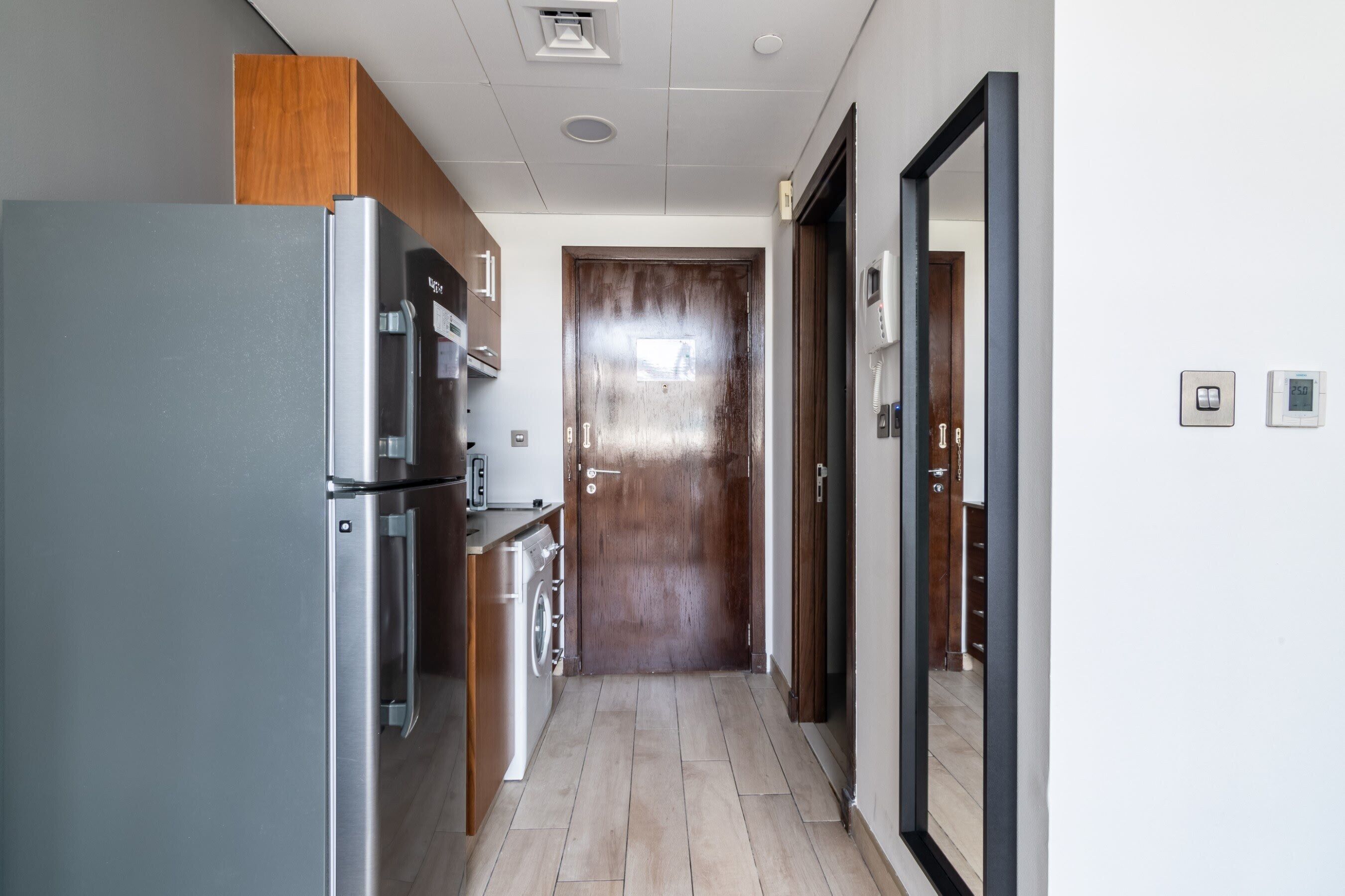 A narrow kitchen with a stainless steel refrigerator, wooden cabinets, a washing machine, and a wooden door at the end.