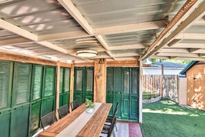 A covered patio with a wooden table and chairs, green shutters, and a small garden area.