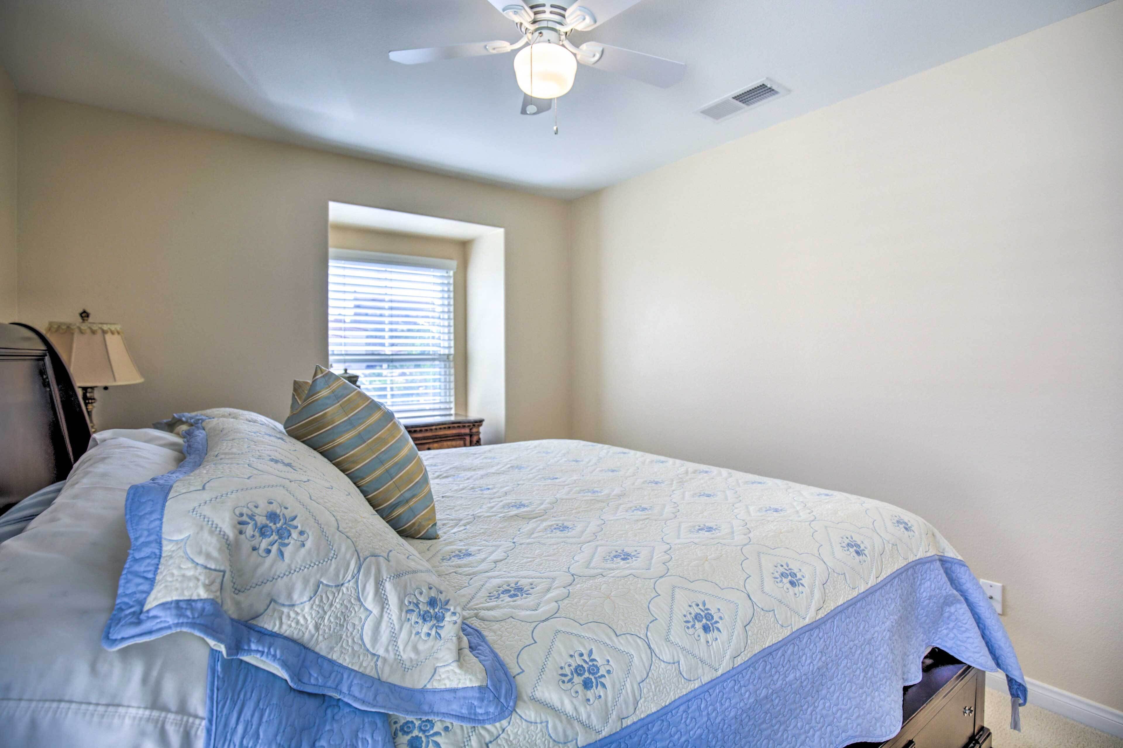 Une chambre à coucher comprenant un lit, un ventilateur de plafond, une fenêtre avec des stores et une table de chevet avec une lampe.