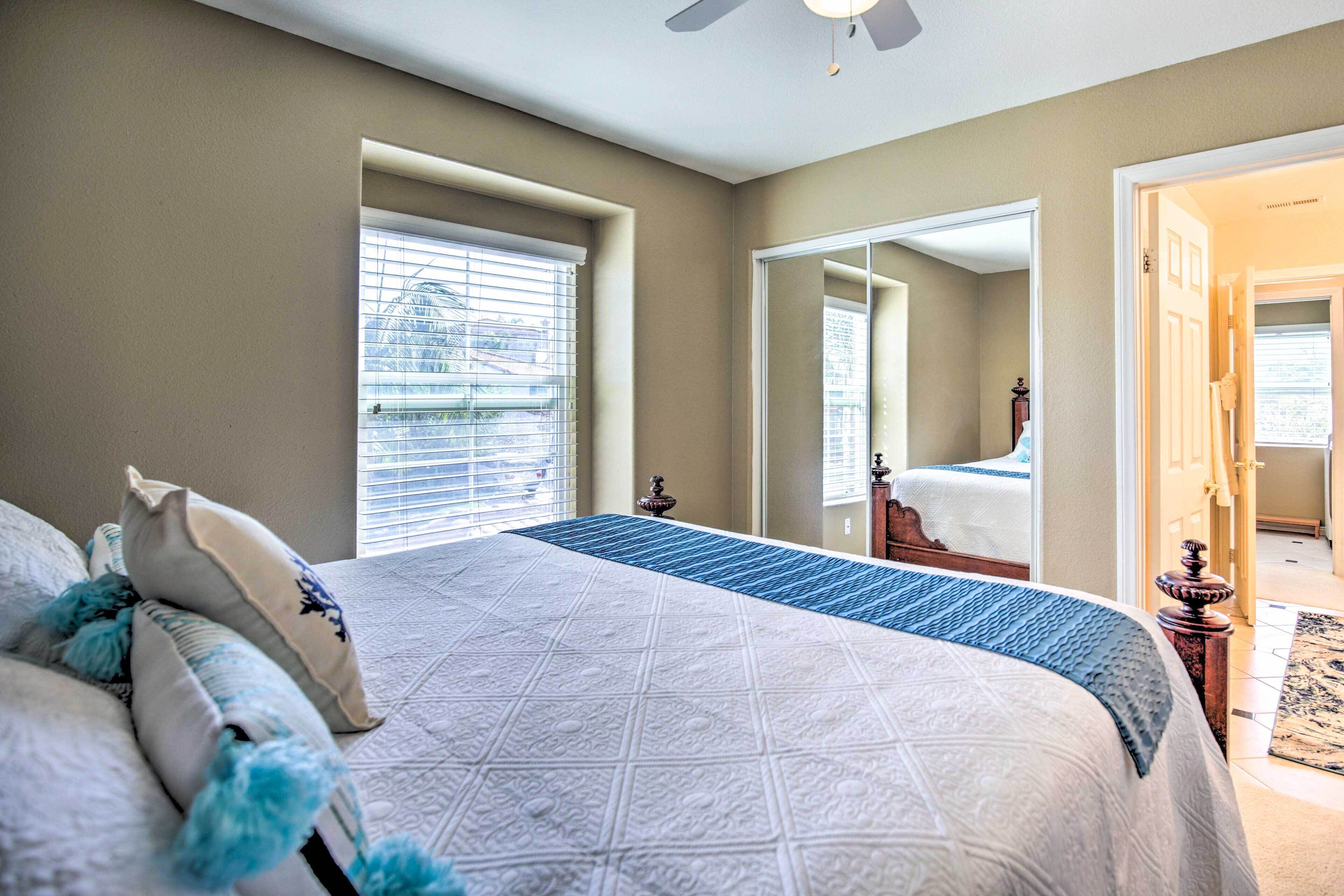 A bedroom with a large bed, a window with blinds, a mirror, and a door leading to another room.
