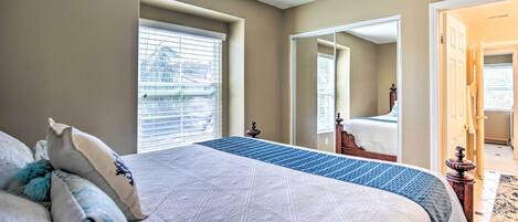 A bedroom with a large bed, a window with blinds, a mirror, and a door leading to another room.