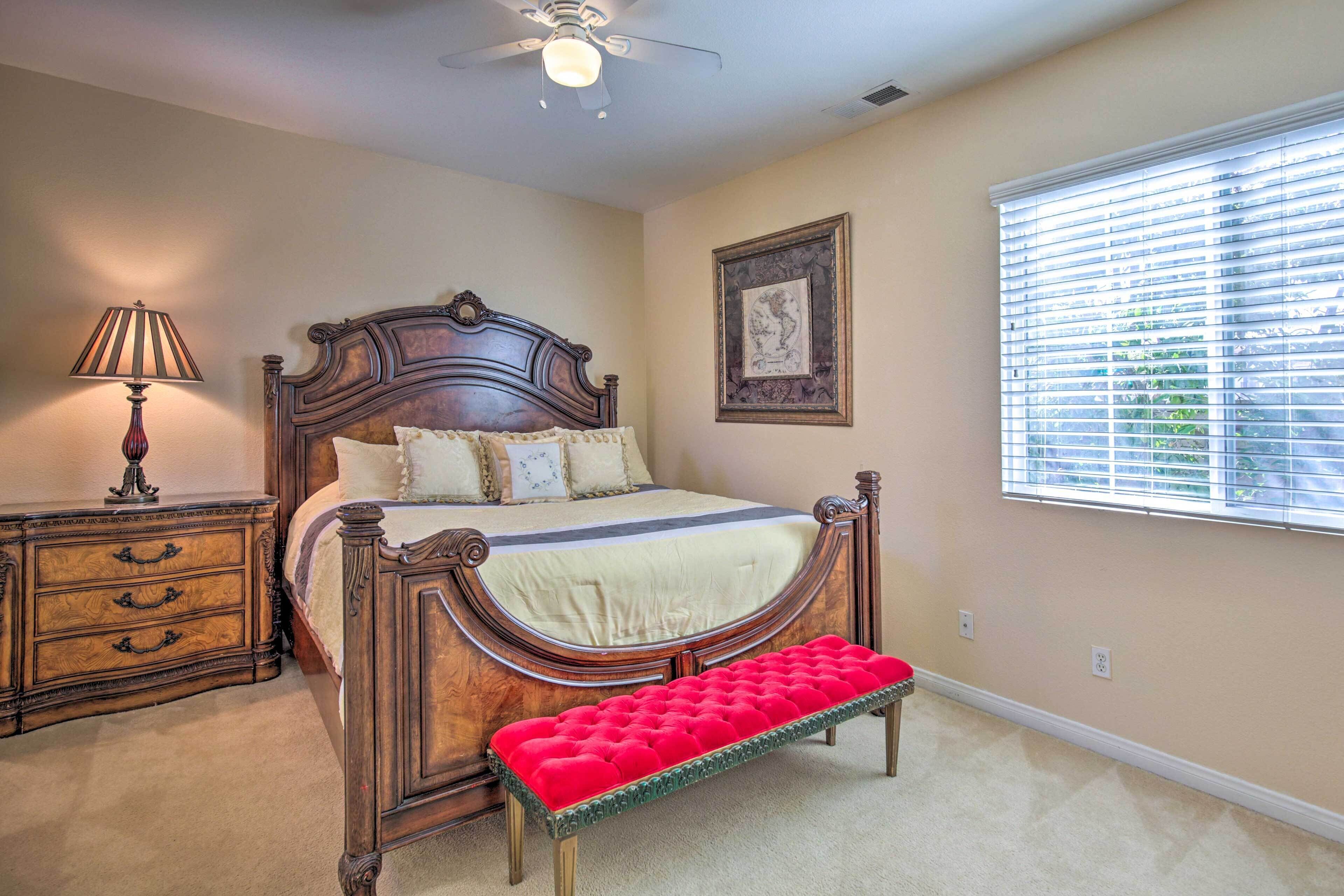 A bedroom with a wooden bed, a bedside table with drawers, a red bench, a ceiling fan, a framed picture on the wall, and a window with blinds.