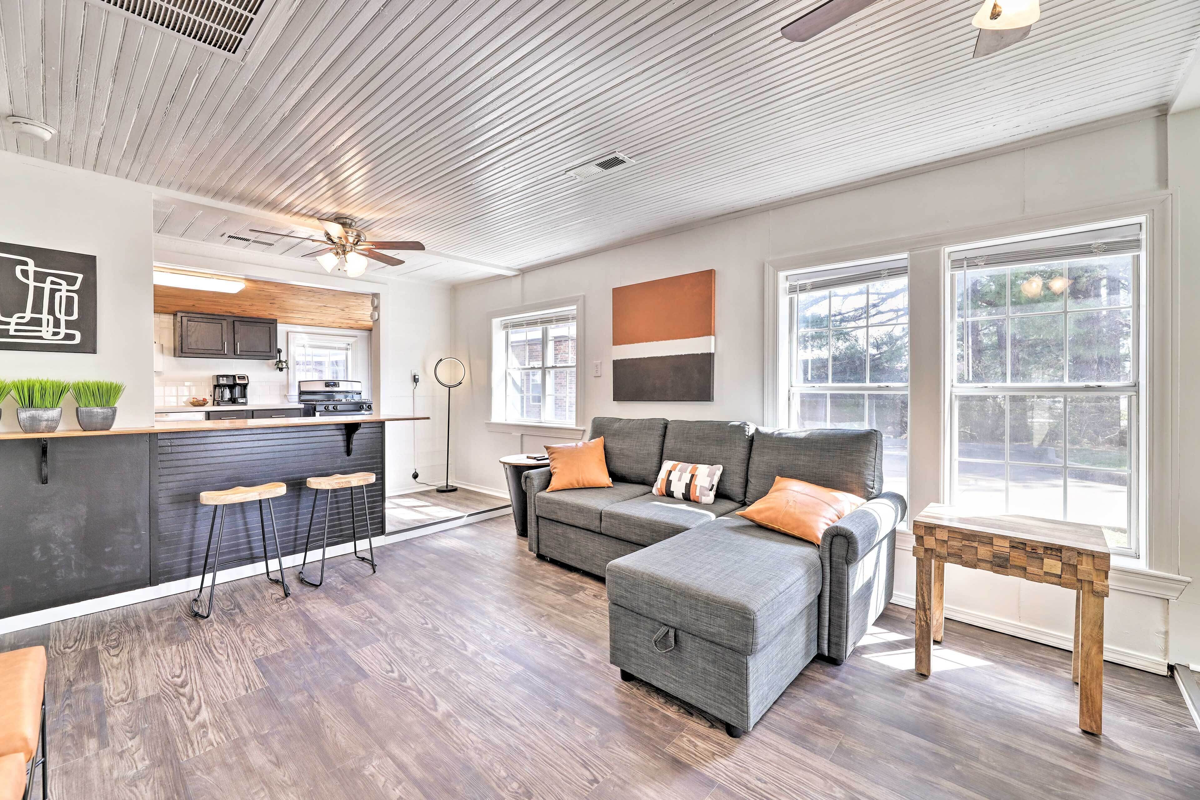 A modern living room with a gray sectional sofa, a wooden coffee table, and a kitchen area with a bar counter and stools.