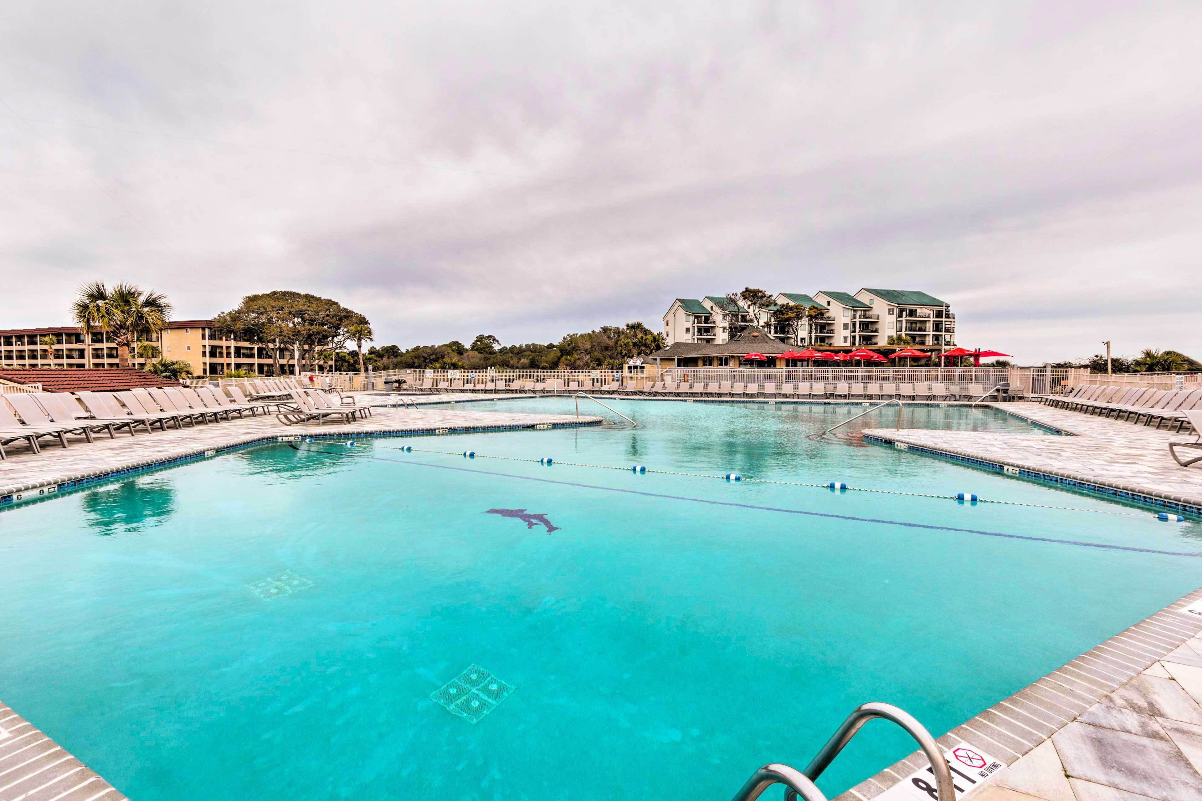 A large outdoor swimming pool with multiple lanes, surrounded by lounge chairs and umbrellas, with buildings in the background.