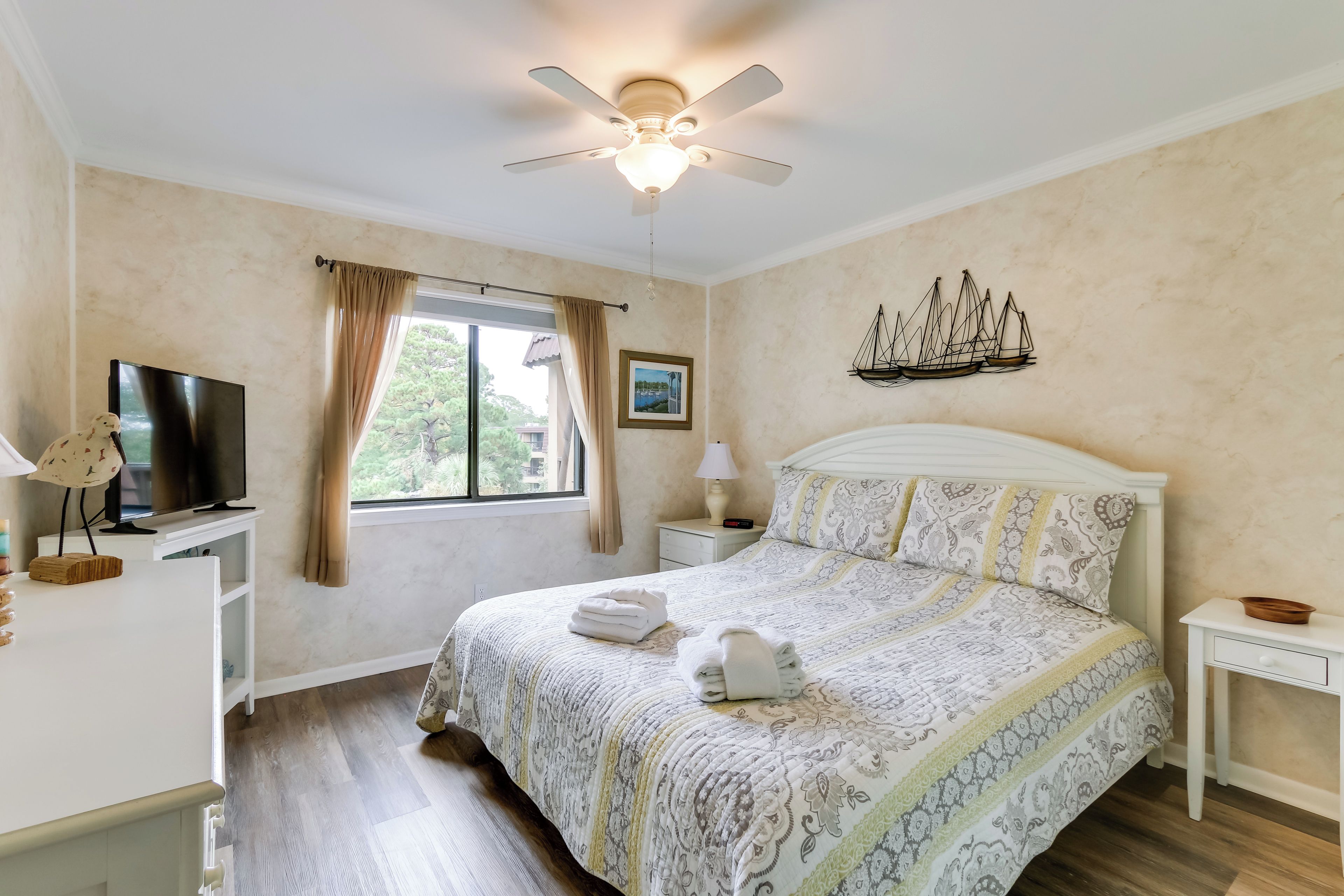 A bedroom with a bed, a television, a ceiling fan, a window with curtains, a nightstand, and a wall decoration.