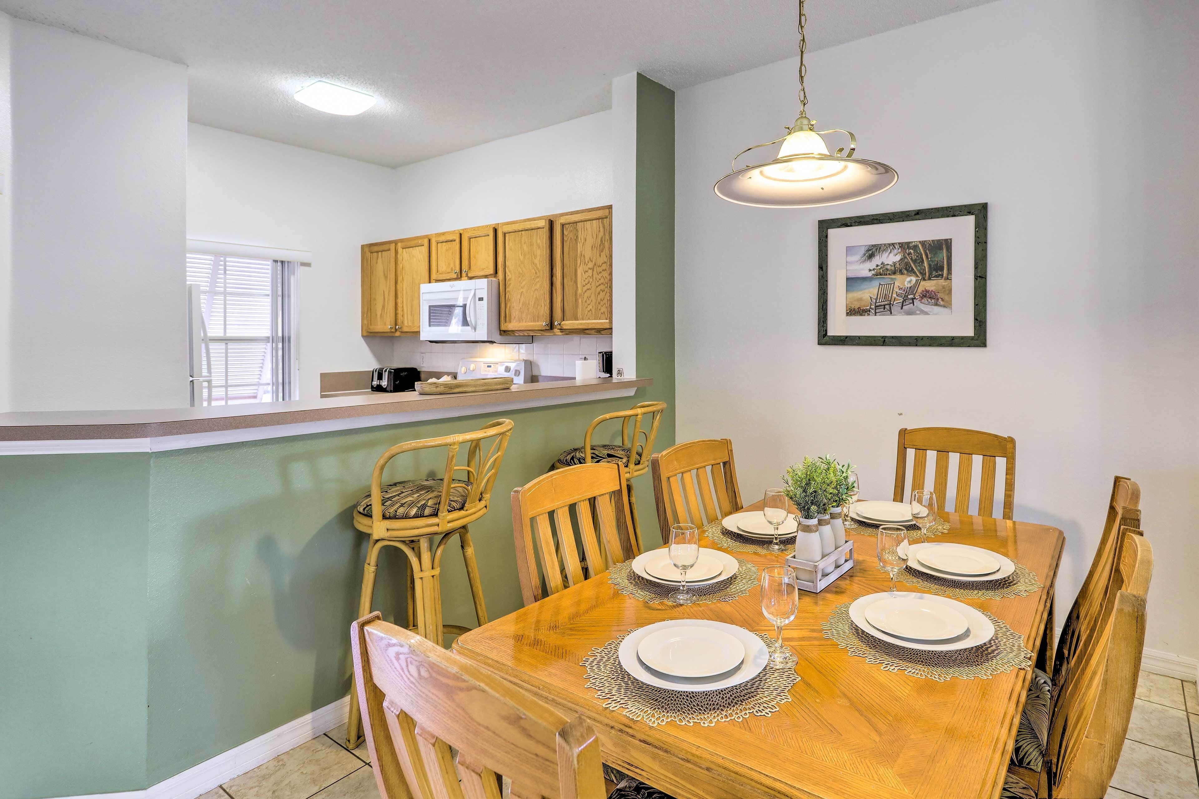 A dining area with a wooden table set for four, surrounded by wooden chairs. A kitchen with wooden cabinets and a microwave is visible in the background.