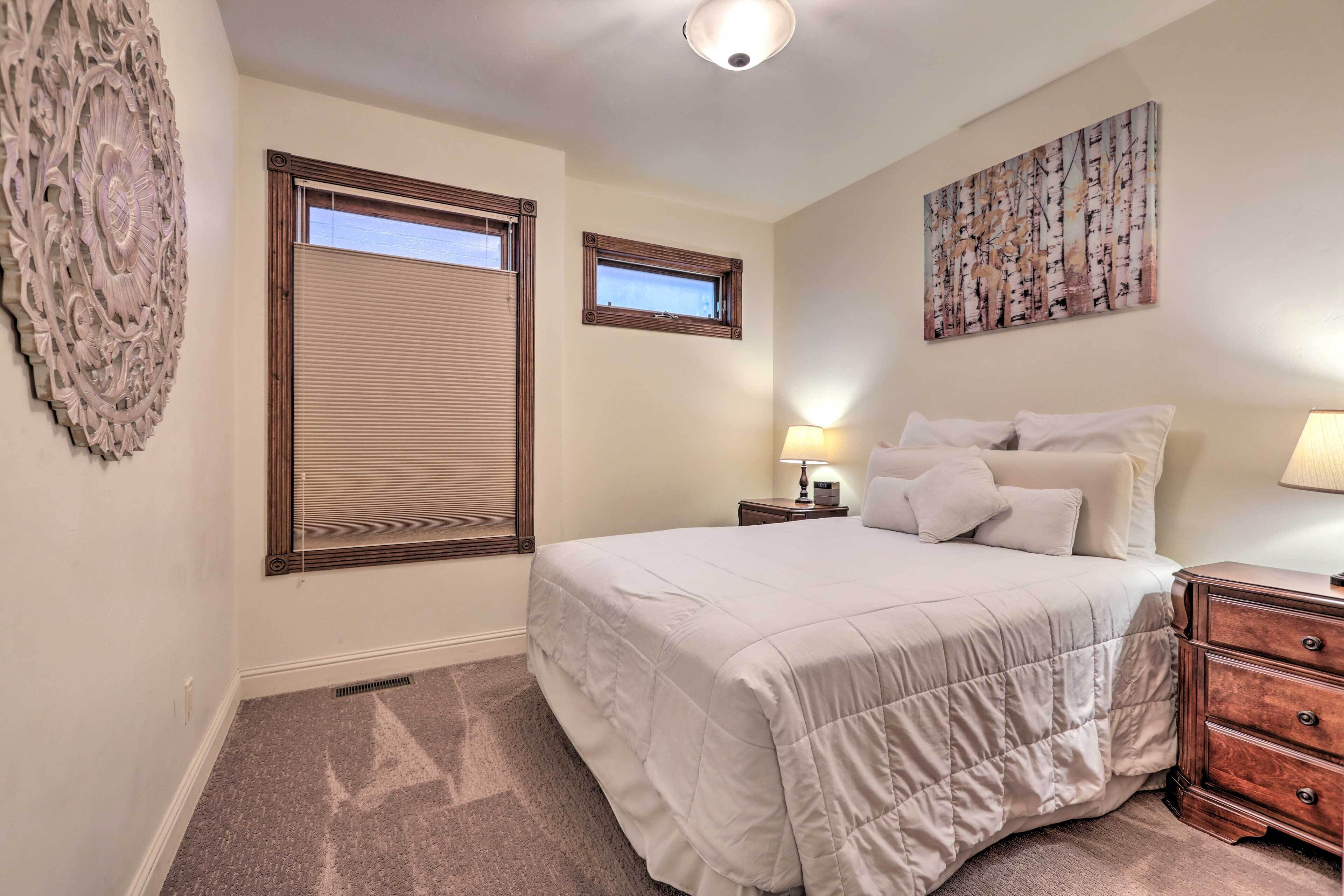 A bedroom with a large bed, a nightstand with a lamp, a dresser, a window with blinds, and a wall art piece.