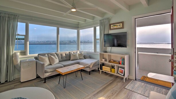 A living room with a sofa, coffee table, and TV, large windows with a view of the water, and a door leading to a balcony. - Chic Bay View Condo < 10 Miles to Dtwn San Diego! (San Diego)