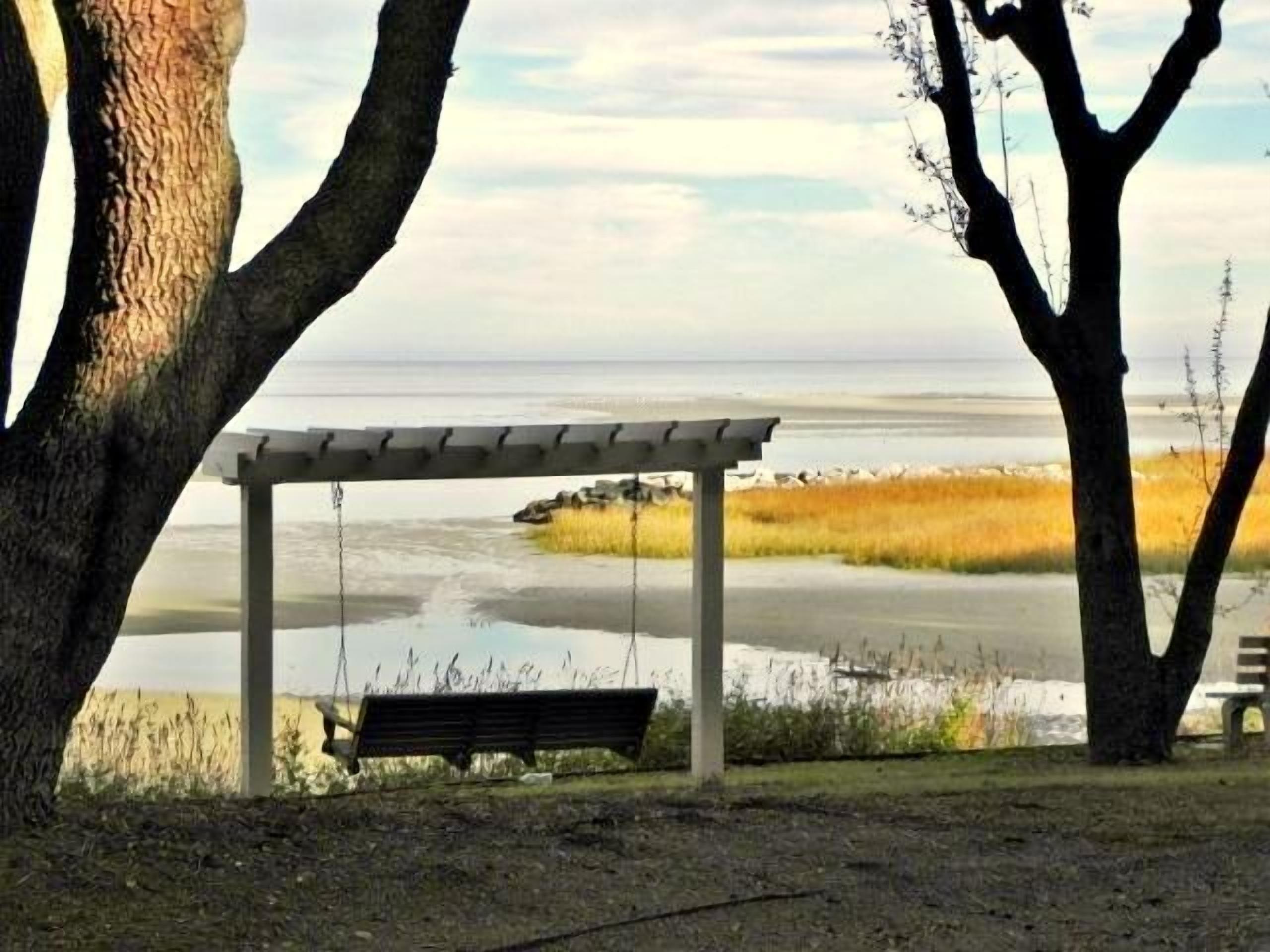 A wooden swing under a pergola overlooking a beach with sand and water.