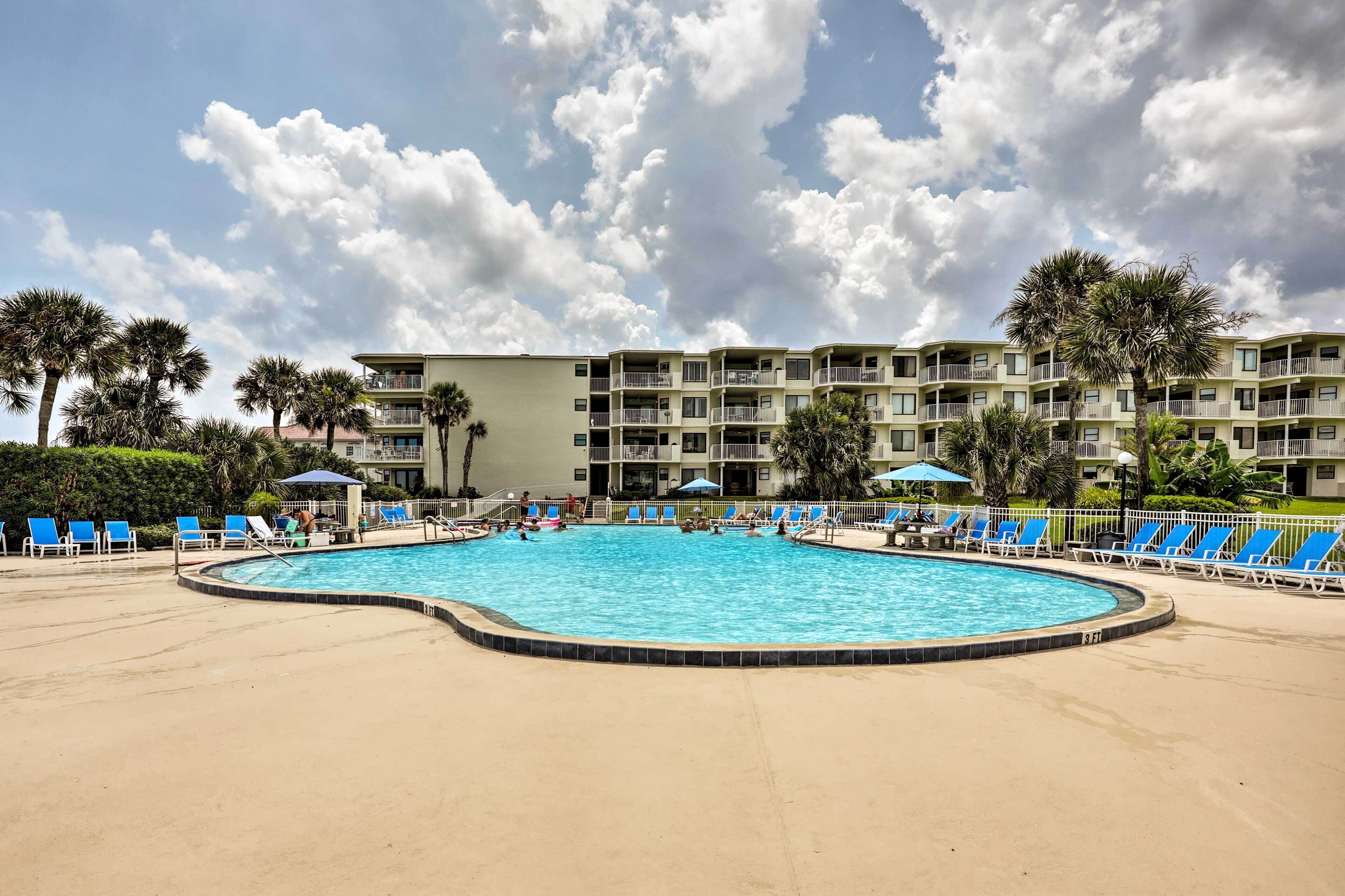 A swimming pool with blue lounge chairs and umbrellas, surrounded by a multi-story building with balconies.