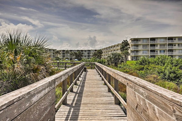 A wooden boardwalk leading to a multi-story building with balconies, surrounded by greenery.