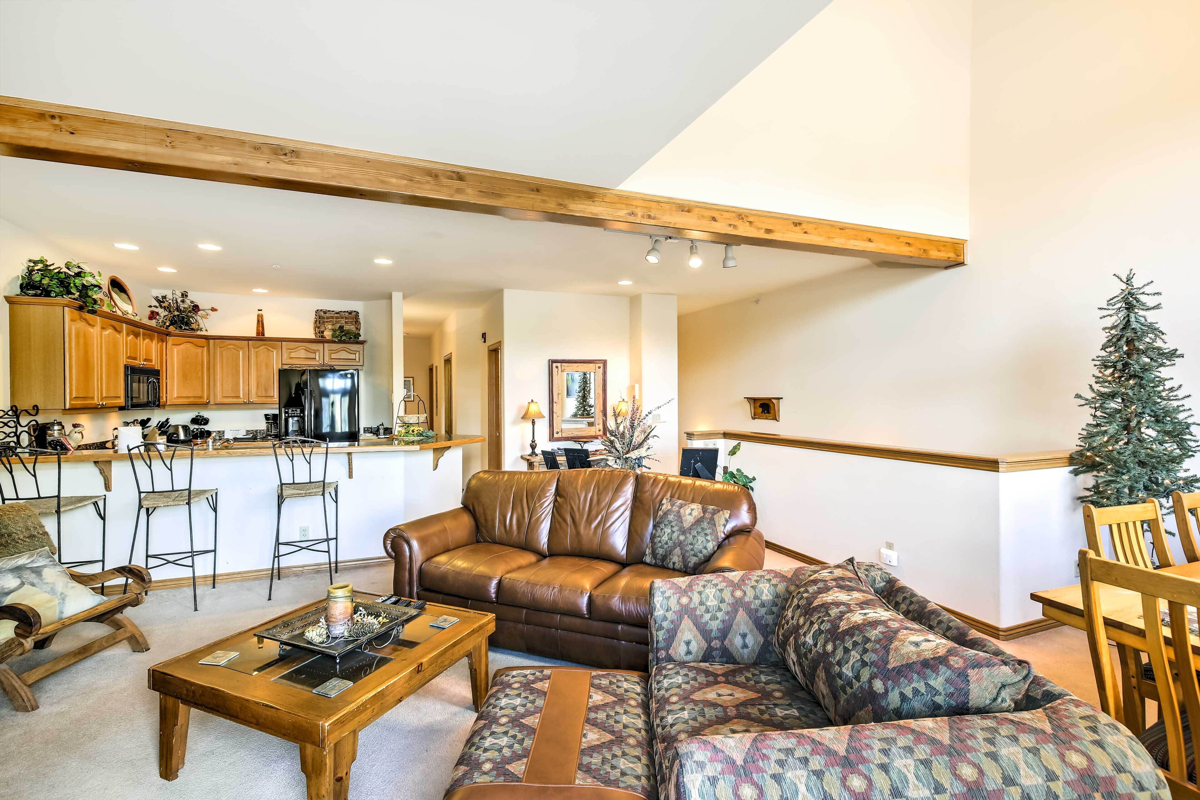 A spacious living area with a brown leather sofa, patterned ottoman, and a wooden coffee table. A kitchen with wooden cabinets and a bar area is visible in the background.