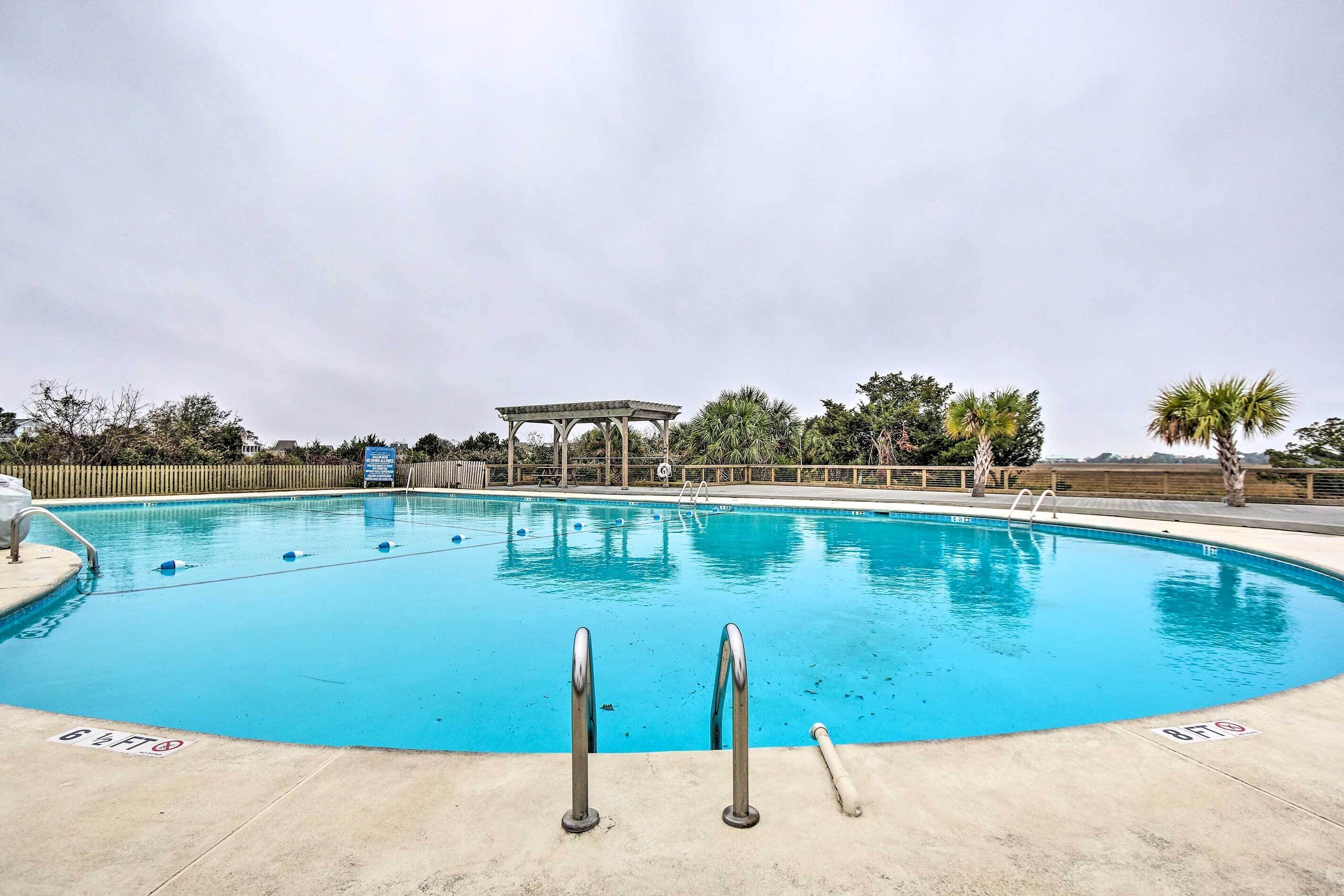 A clear blue swimming pool with two metal ladders in the foreground, surrounded by palm trees and a wooden fence.