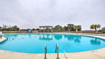 A clear blue swimming pool with two metal ladders in the foreground, surrounded by palm trees and a wooden fence.