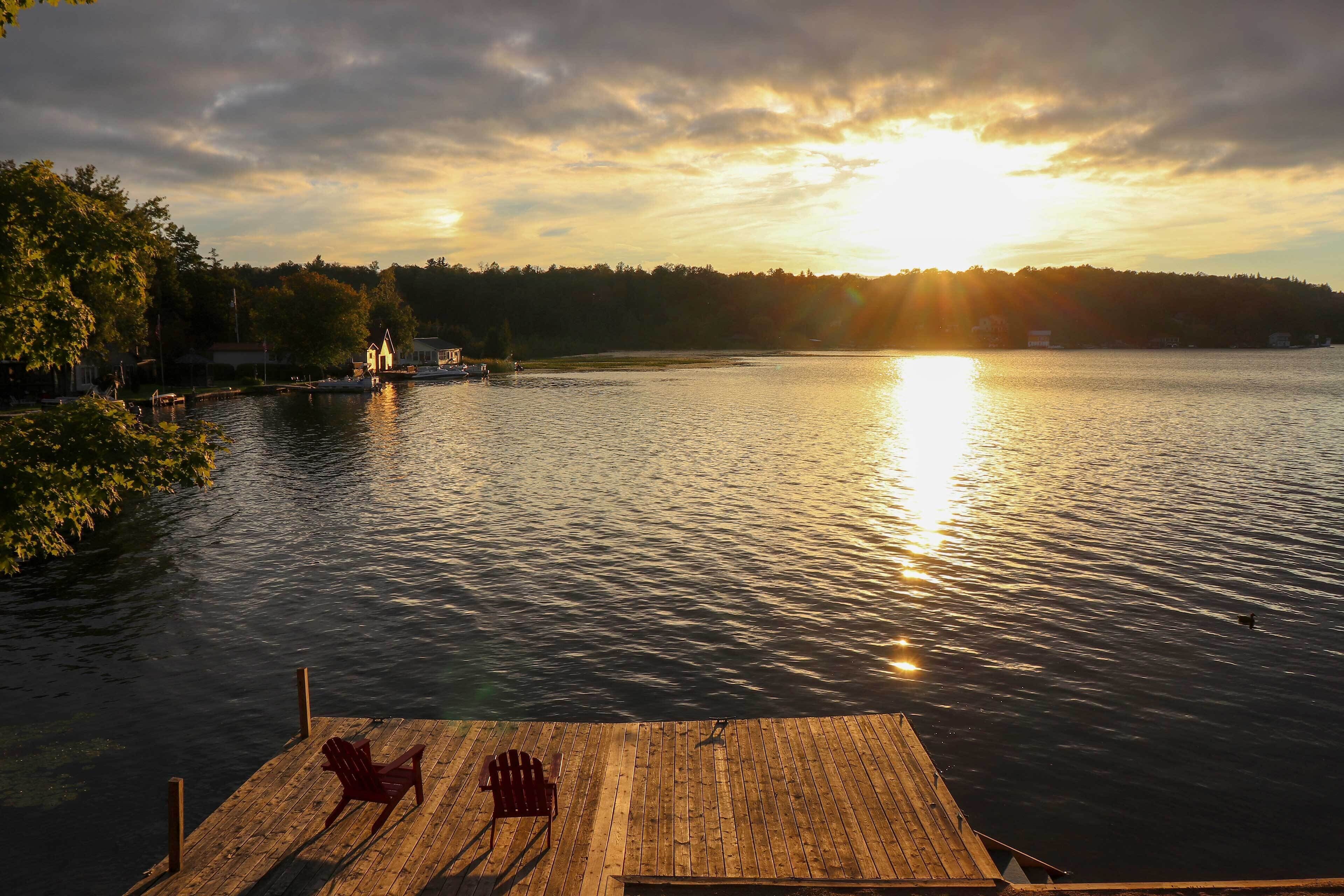 A wooden dock with two chairs, a calm lake, and a sunset in the background.