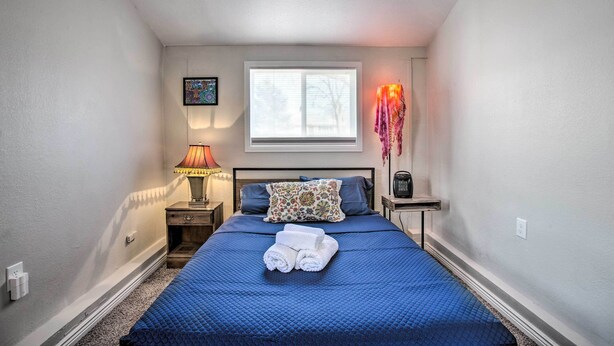 Een net opgemaakt bed met blauw beddengoed, een bedlampje, een klein tafeltje met een lamp en een raam met jaloezieën.