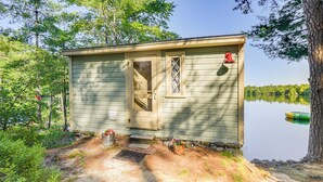 A small, single-story cabin with a door and window, situated by a lake with trees in the background.