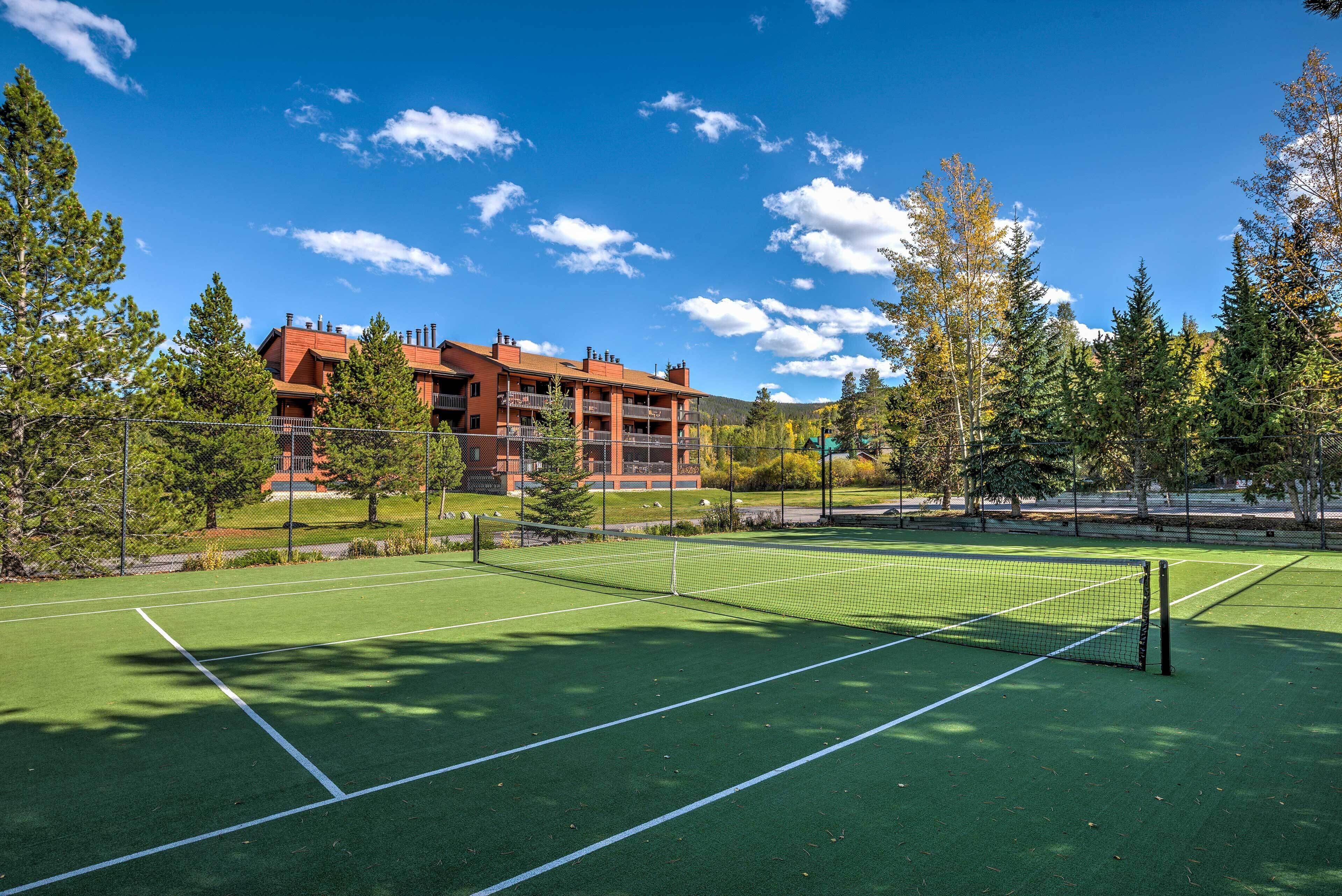A tennis court with a net, surrounded by trees and a building in the background.