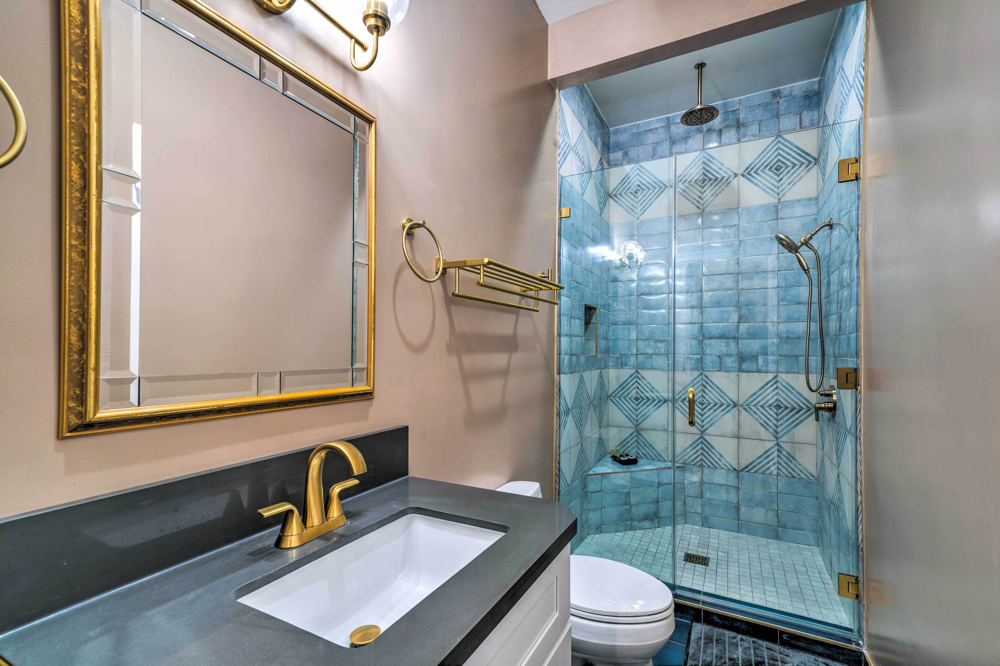 A bathroom with a gold-framed mirror, a black countertop, a white sink, and a glass-enclosed shower with blue patterned tiles.