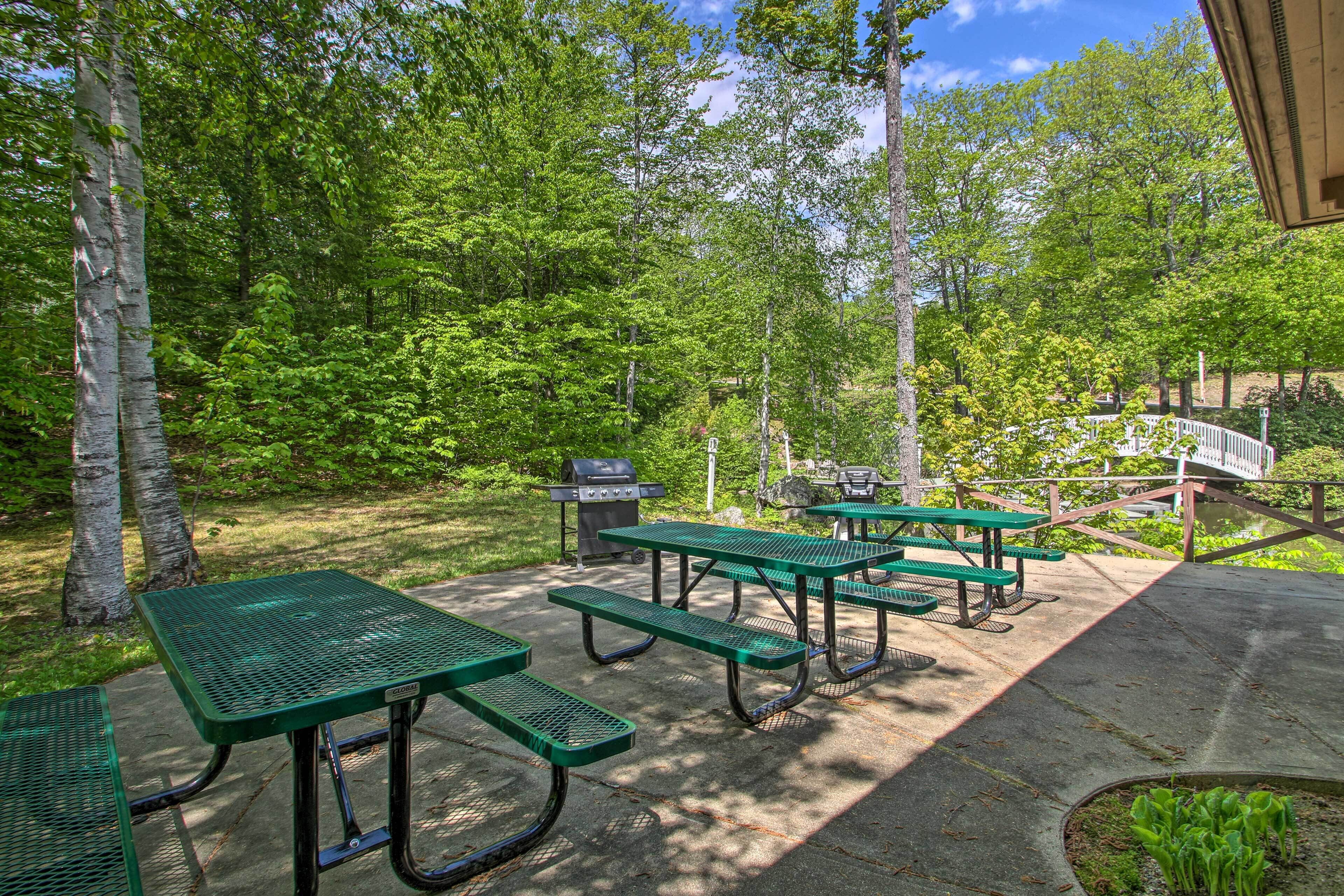 A picnic area with green metal tables and benches, surrounded by trees and a grill.