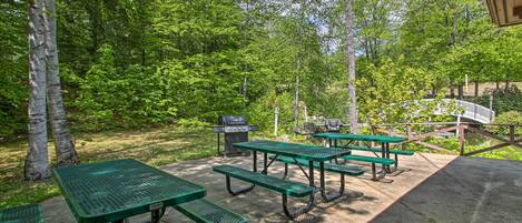 A picnic area with green metal tables and benches, surrounded by trees and a grill.
