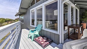 A covered deck with a white railing, a turquoise chair, a wooden table, and a large window overlooking a landscape. - Irish Beach Cottage w/ Beach Access & Ocean Views (Manchester)