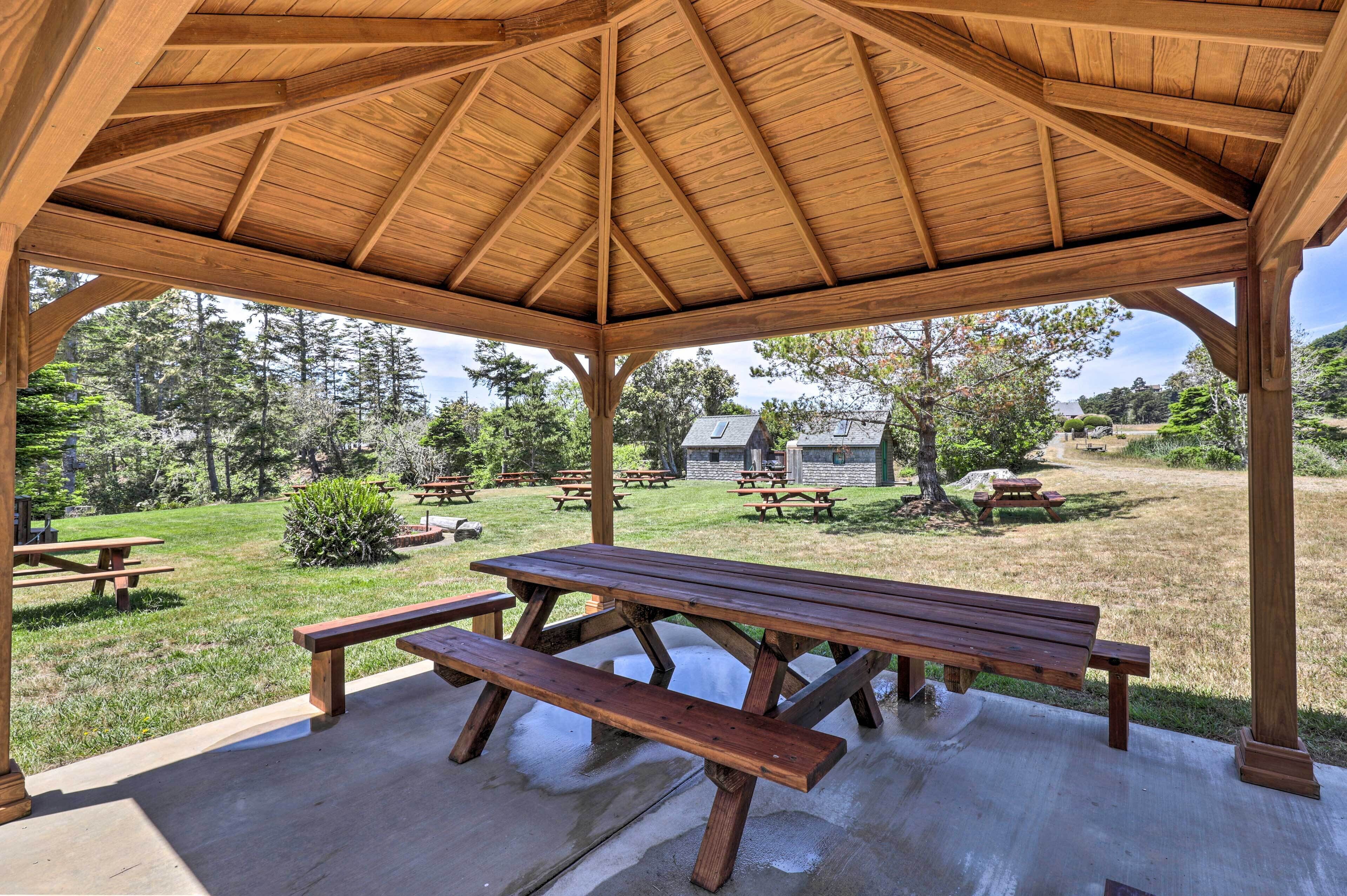 A covered picnic area with wooden benches and picnic tables, surrounded by a grassy landscape and trees.