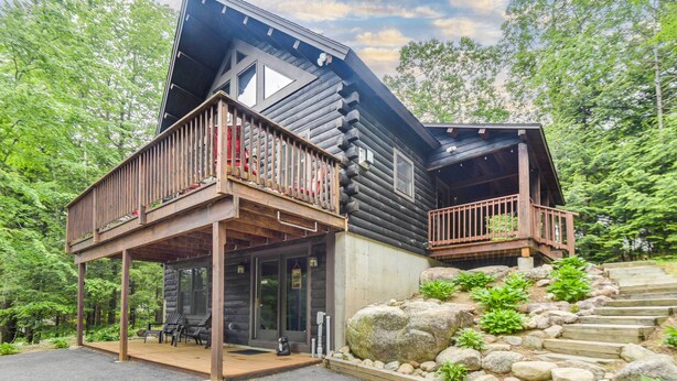A log cabin with a wooden deck, a stone foundation, and a staircase leading up to a second level. - Updated Log Cabin Near Story Land & Diana's Baths! (Conway)